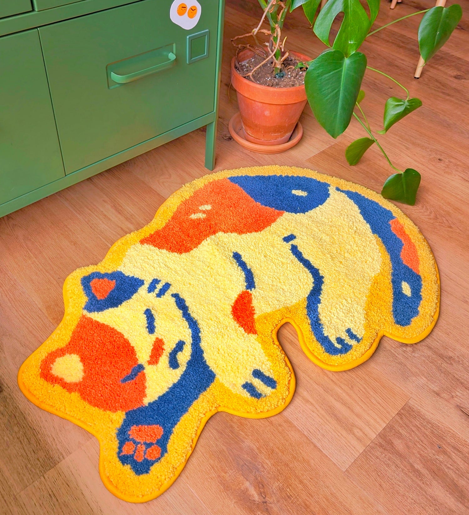 Colorful cat-shaped rug on a wooden floor with a green cabinet and plant in the background.